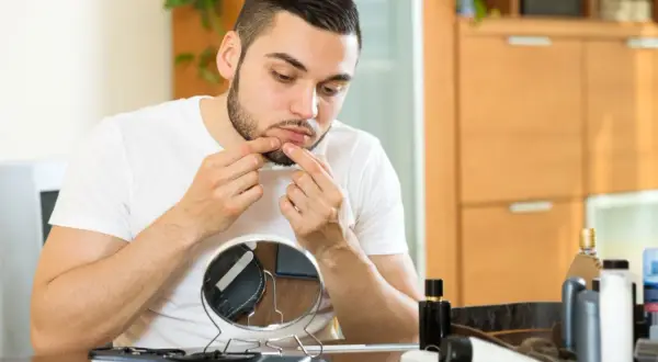 Mężczyzna ogląda pryszcza w lusterku i dotyka swojej twarzy.
